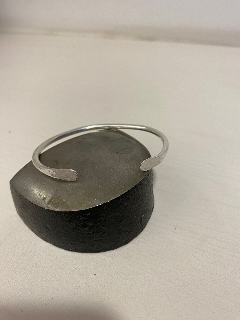 En enkel silverfärgad armring ligger på ett halvt klotformat metallblock på ett vitt underlag.