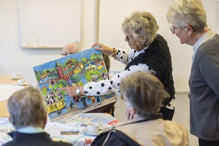 En grupp äldre personer sitter runt ett bord och tittar på en färgglad målning som en kvinna håller upp.