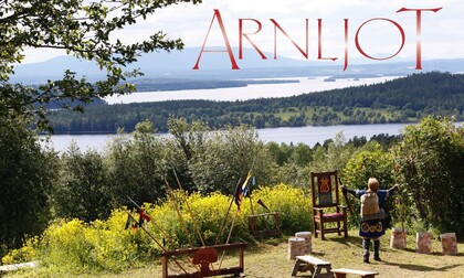 Utsikt över sjö och skog med en person i historisk dräkt, stolar och flaggor, texten 'ARNLJOT' syns.