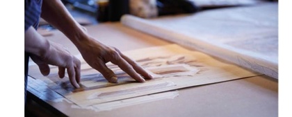 Två händer arbetar med ritningar eller mönster på ett bord i ett ljust rum.