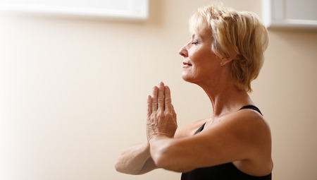 Äldre kvinna med kort blont hår gör yogaposition med händerna framför bröstet och ögonen slutna.