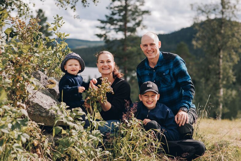 En familj på fyra sitter tillsammans utomhus i naturen bland gräs och buskar, med skog och berg i bakgrunden.