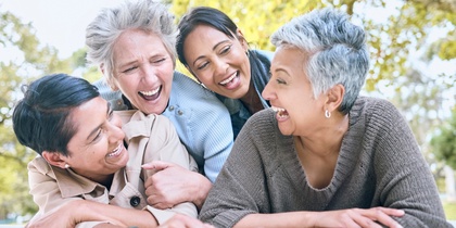 Fyra glada kvinnor i medelåldern skrattar tillsammans utomhus en solig dag.