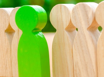 En grön träfigur står mellan två rader av ljusa träfigurer på ett bord med grön bakgrund.