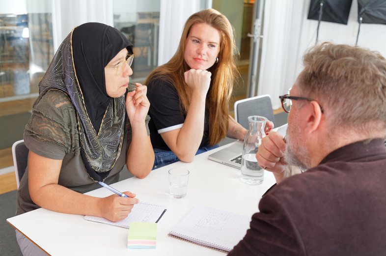 Tre personer sitter vid ett bord och diskuterar, med anteckningsblock och glas vatten framför sig.