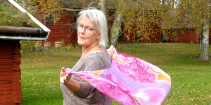 Äldre kvinna med glasögon står utomhus och håller en rosa sjal som blåser i vinden.