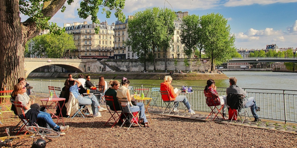 Folk sitter på uteservering vid floden med utsikt över bro, träd och klassiska byggnader i solsken.