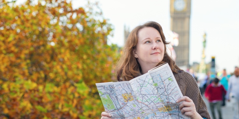 Kvinna håller en karta och står utomhus nära Big Ben i London, med höstlöv i bakgrunden.