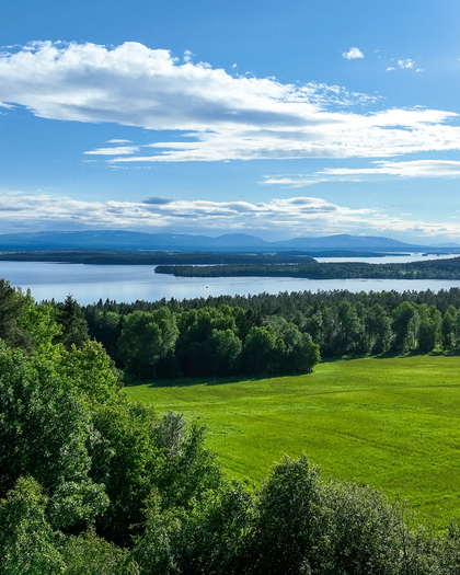 Landskapsvy med gröna ängar, skog, en sjö och blå himmel med moln i bakgrunden.