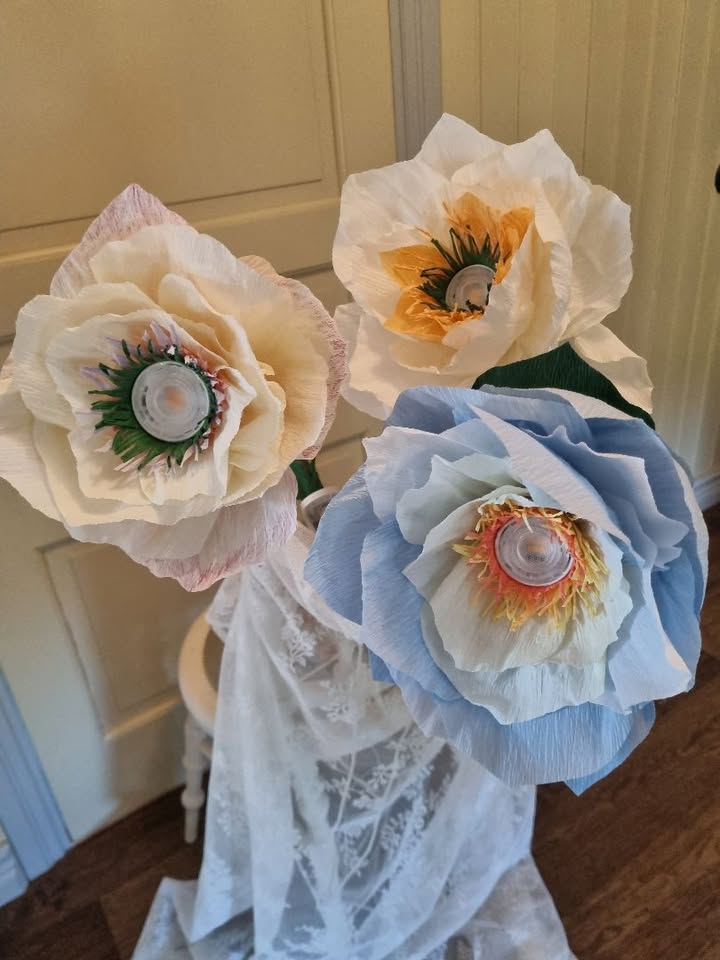 Tre stora konstgjorda blommor av papper med plastlock i mitten, i pastellfärger, står mot en ljus bakgrund.