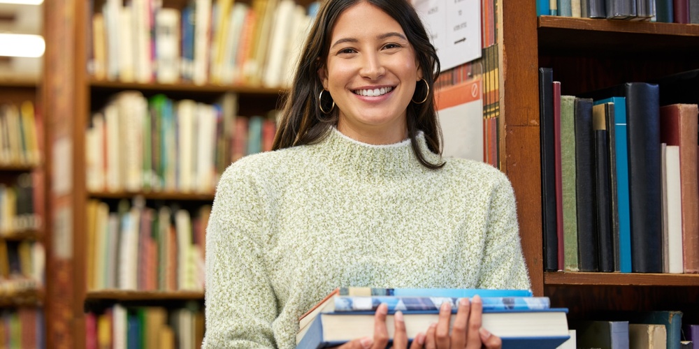 Ung kvinna med långt mörkt hår håller böcker och ler i ett bibliotek.