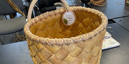 En handflätad korg av naturmaterial står på ett svart bord i ett rum med stolar och hantverksmaterial.