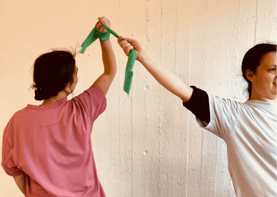 Två personer håller i ett grönt band och sträcker upp armarna mot varandra framför en ljus vägg.