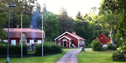 En grusväg leder till tre röda hus omgivna av grönska och träd, med flaggstänger och en grillplats.