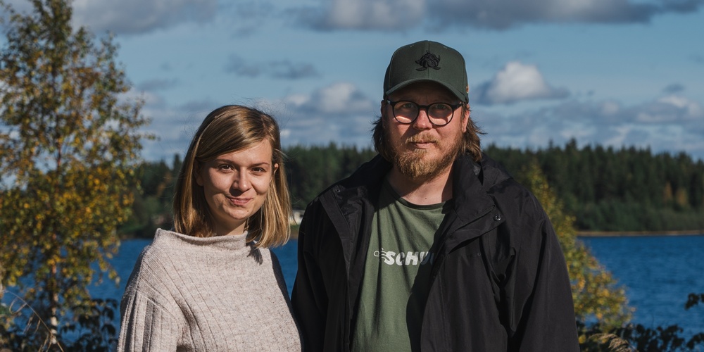 En kvinna och en man står utomhus vid en sjö med skog i bakgrunden under en solig dag.