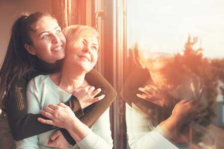 En leende kvinna kramar om en annan kvinna bakifrån framför ett fönster.