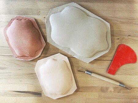 Tre formade lerkupor, ett rött plastverktyg och ett träsnidad lerverktyg på ett träbord.