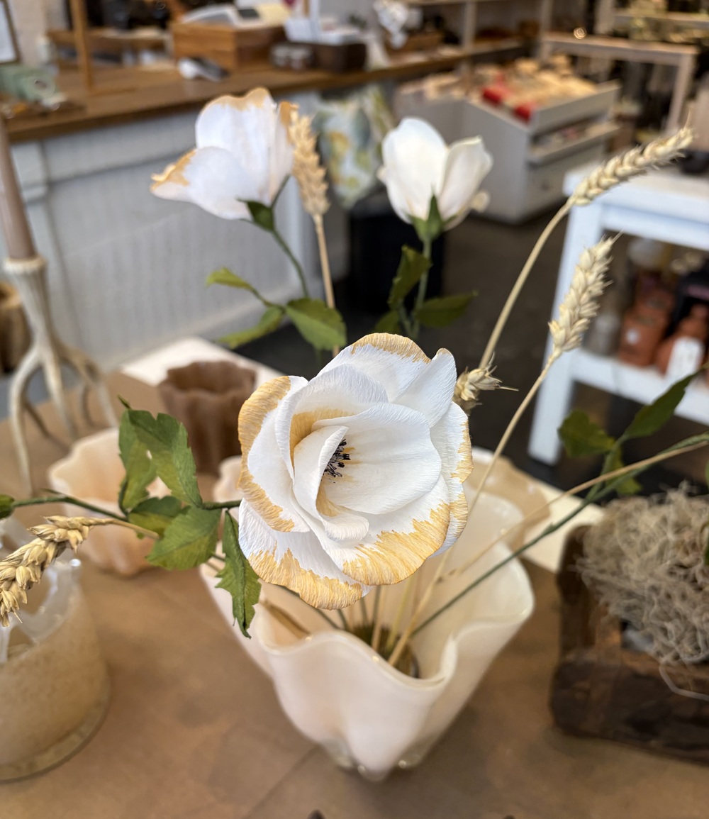 Vita konstgjorda blommor med gula kanter i en vit vas på ett bord i en butiksmiljö.