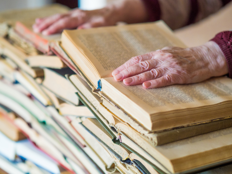 Två händer bläddrar i en gammal bok ovanpå en hög med andra böcker.