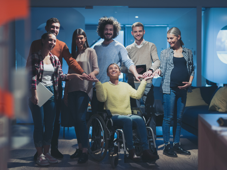 En grupp glada människor står runt en kvinna i rullstol och håller hennes händer inomhus.