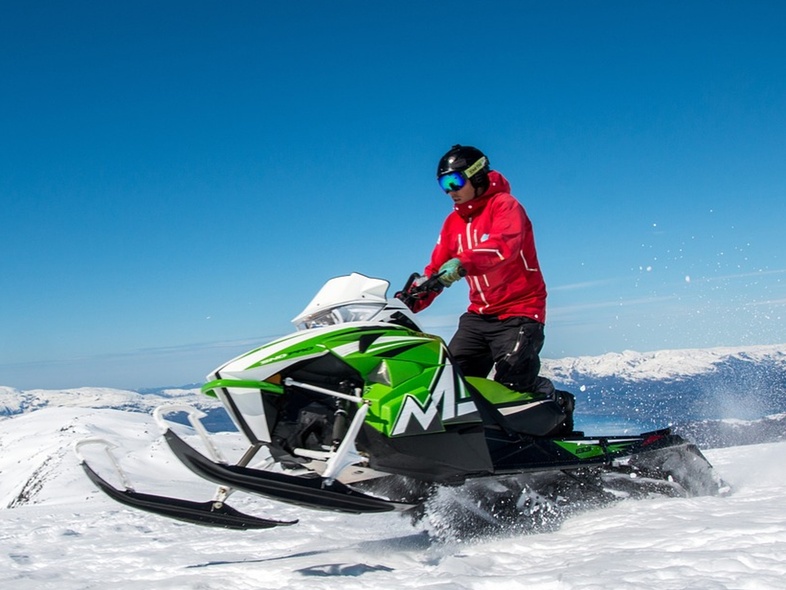 Person kör en grön snöskoter på ett snötäckt fjäll under klarblå himmel.