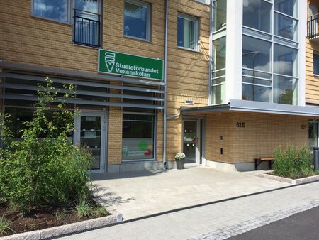 Entré till en modern byggnad med skylt för Studieförbundet Vuxenskolan, omgiven av växter och gångväg.