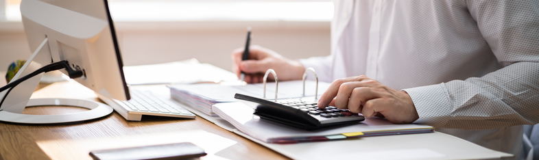 Person arbetar vid skrivbord med dator, pärm och miniräknare, skriver anteckningar.