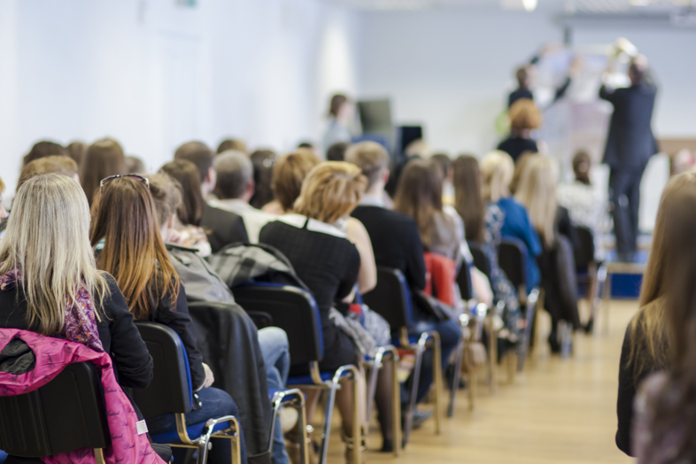 En mängd personer sitter i en aula med ryggen mot kameran och lyssnar på vad två personer presenterar på scenen.