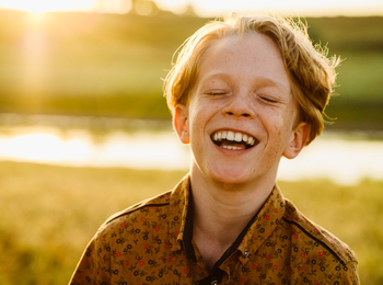 En ung pojke skrattar glatt utomhus i solnedgången med grönt landskap i bakgrunden.