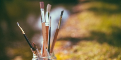 Målarpenslar i en glasburk, två färgburkar och en liten trädocka utomhus på gräs vid vatten.