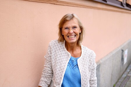 Leende äldre kvinna med glasögon står mot en ljusrosa vägg utomhus.