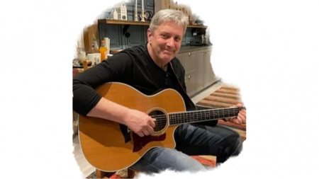 Man sitter inomhus och spelar akustisk gitarr, ler mot kameran.