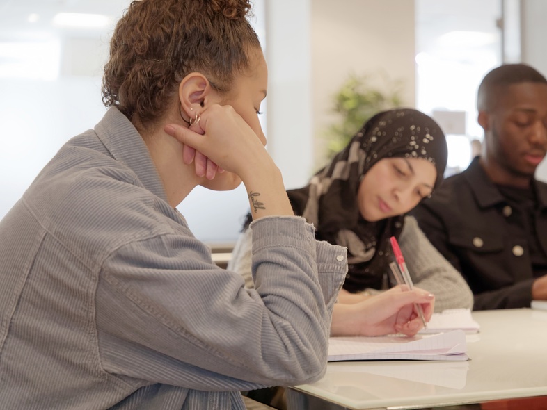 Tre unga personer sitter vid ett bord och studerar tillsammans, en skriver i ett block.