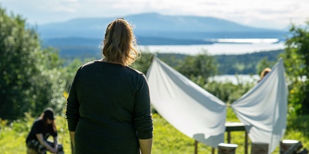 En person står på en gräsmatta och tittar mot två andra vid ett vitt tyg utomhus, med berg och sjö i bakgrunden.