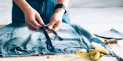 Person arbetar med att sy om ett par jeans, med dragkedja, måttband och sytråd på bordet.