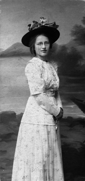 Svartvit foto av kvinna i långklänning och stor hatt, poserande framför målad bakgrund.