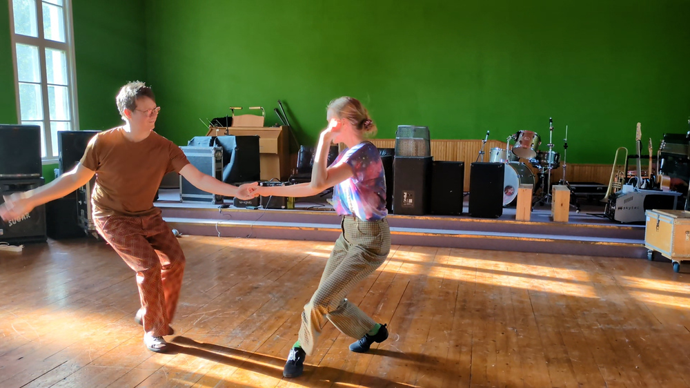 Två personer dansar i ett solbelyst rum med trägolv och musikutrustning i bakgrunden.