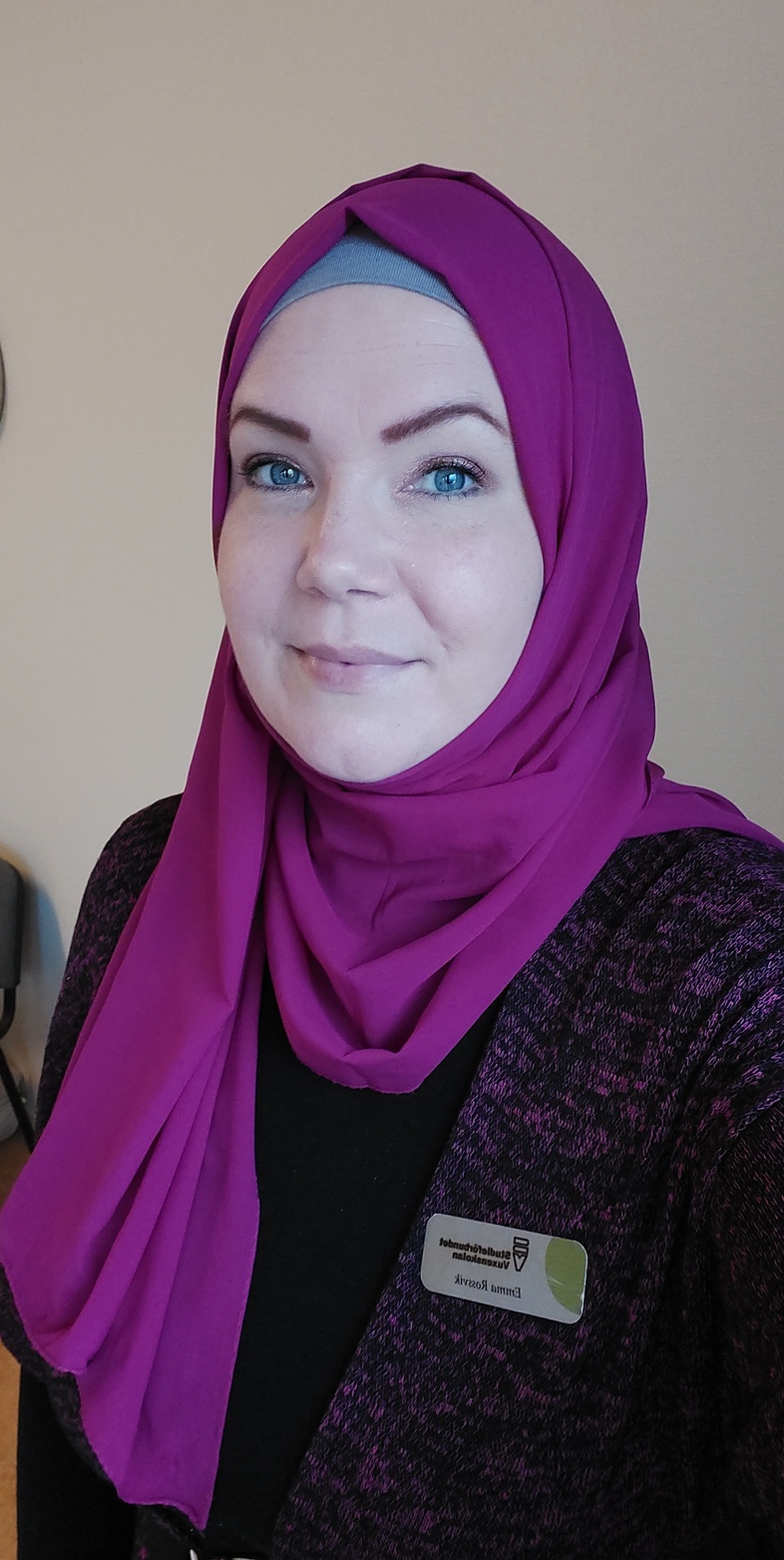 Emma Rossvik med lila hijab och namnskylt, ler mot kameran i inomhusmiljö.
