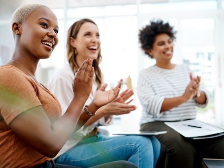 Tre kvinnor sitter tillsammans inomhus och applåderar, ser glada och engagerade ut.