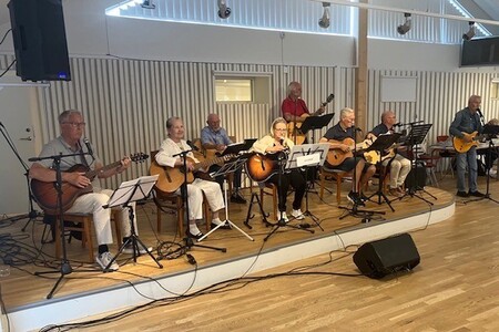 En grupp äldre personer spelar gitarr och sjunger på en scen i en ljus lokal.