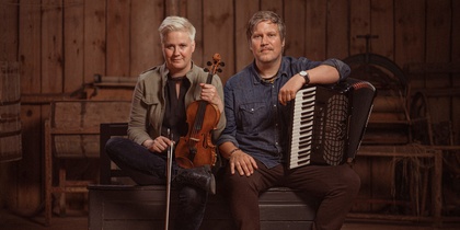 Två personer sitter på en bänk i en trästuga, en håller en fiol och den andra ett dragspel.