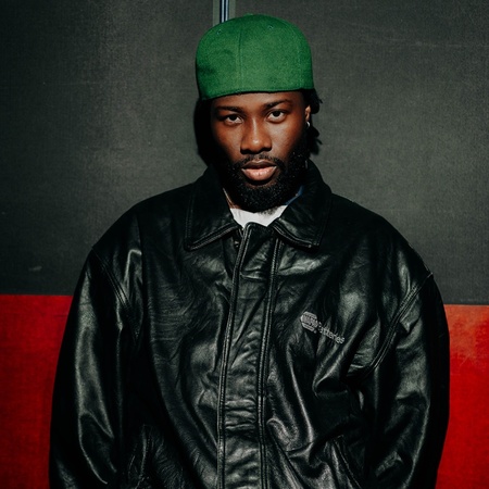 Mbo Momo med grön mössa och svart läderjacka står mot en svart och röd vägg.