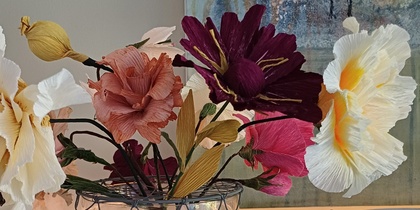 En glasvas med färgglada konstgjorda blommor står på ett turkost bord, med en tavla och dekorationer i bakgrunden.