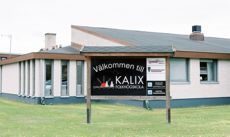 Skylt med texten 'Välkommen till Kalix folkhögskola' framför en låg, ljus byggnad med gräsmatta.