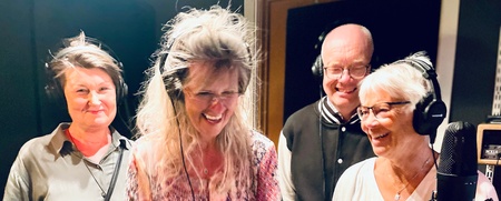 Fyra vuxna personer står tillsammans i en inspelningsstudio, alla bär hörlurar och ler mot mikrofonen.