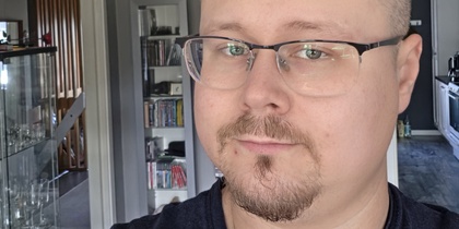 Man med glasögon och kort skägg tar en selfie i ett modernt vardagsrum med grå väggar.