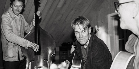 Tre personer spelar akustiska instrument i ett rum med träpanel, omslag till albumet 'Medan Viskan rinner' av Claes Pihl.