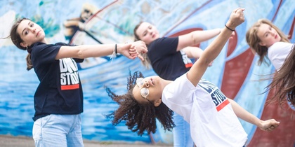 Fyra unga personer dansar energiskt framför en blå graffitivägg utomhus.