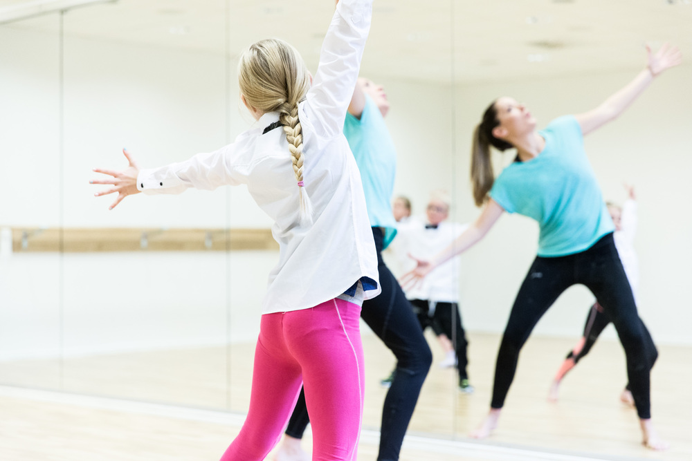 Personer dansar i en ljus dansstudio med spegelvägg, en kvinna i rosa tights i förgrunden.
