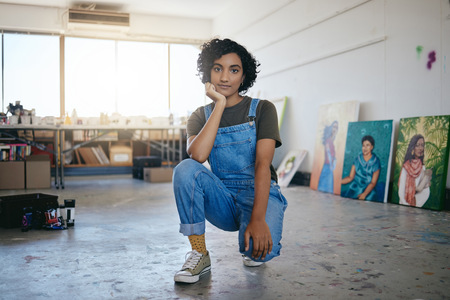 Ung kvinna i blå snickarbyxor sitter på huk i en konststudio med målningar i bakgrunden.